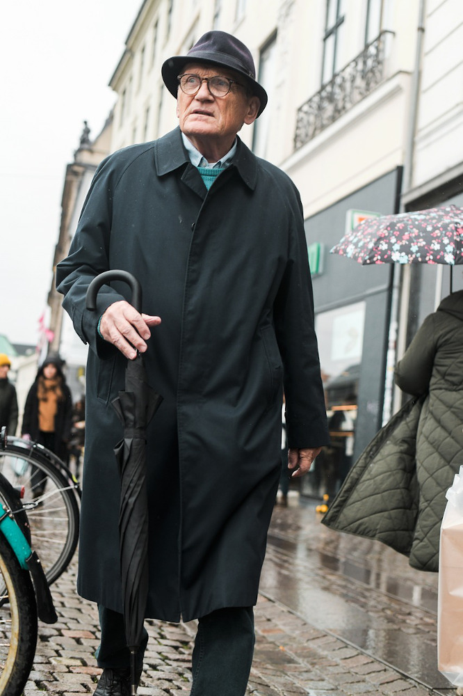 Elderly man in street in coat and hat with umbrella.